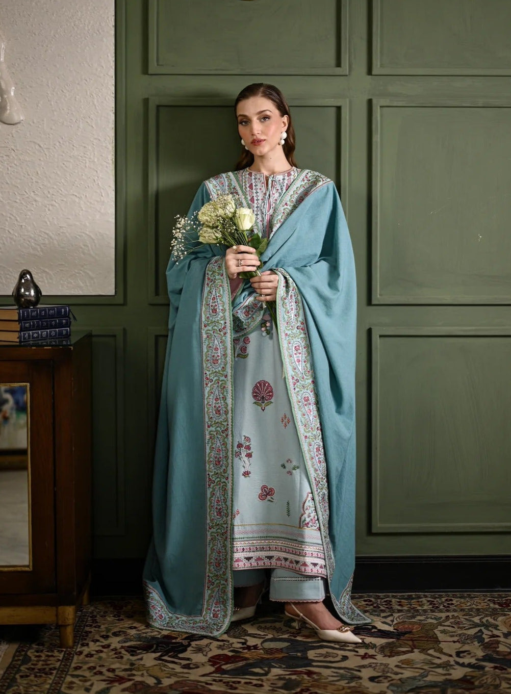 Woman in a light blue traditional outfit holding flowers against a green paneled wall.